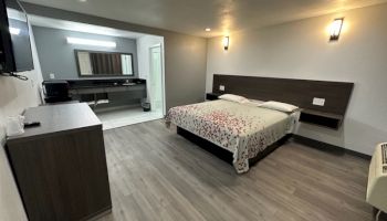 A modern hotel room featuring a bed, bedside tables, a TV, desk, and open bathroom. The decor is minimalistic with warm lighting.