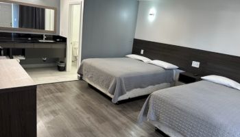 The image shows a modern hotel room with two beds, a bathroom, and a vanity area, featuring a minimalist design and wood flooring.