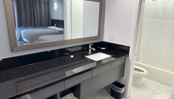 A hotel bathroom with a large mirror, sink, and toiletries on a black countertop. Bedroom visible through doorway, white shower curtain.