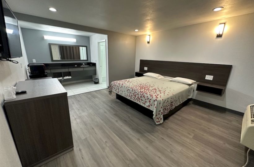 A modern hotel room with a double bed, wooden flooring, a TV, and a desk area. The decor is simple and clean.