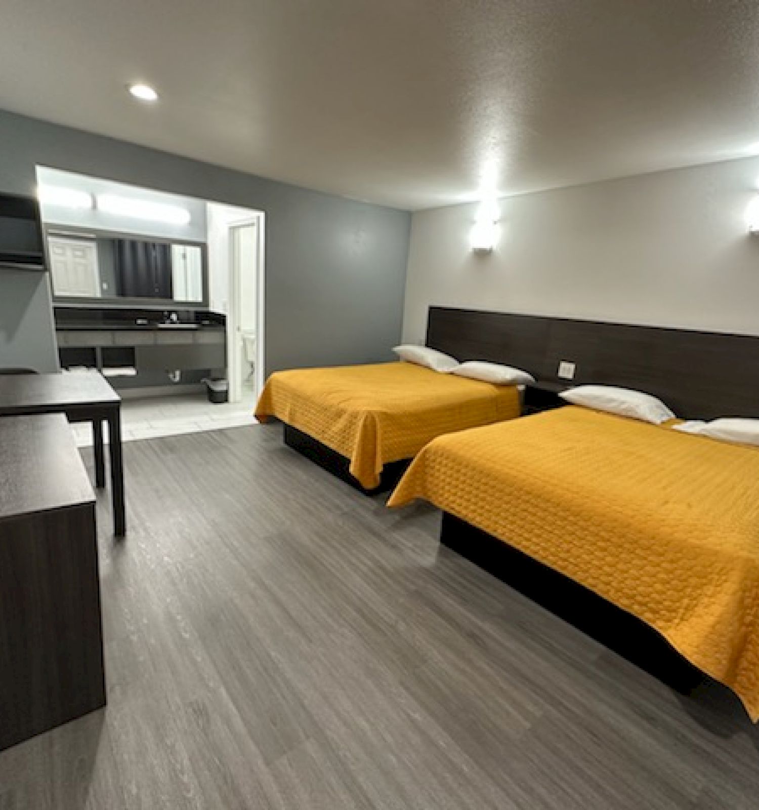 A hotel room features two double beds with yellow covers, a TV, a desk, and a bathroom vanity area visible in the background.