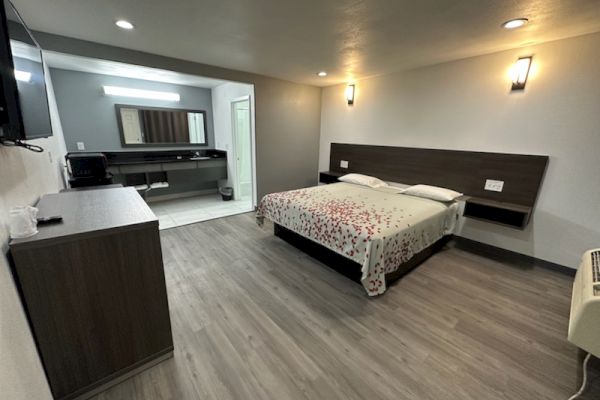 A modern hotel room with a double bed, TV, dresser, and a bathroom in view. The decor is minimalistic with wooden flooring and soft lighting.