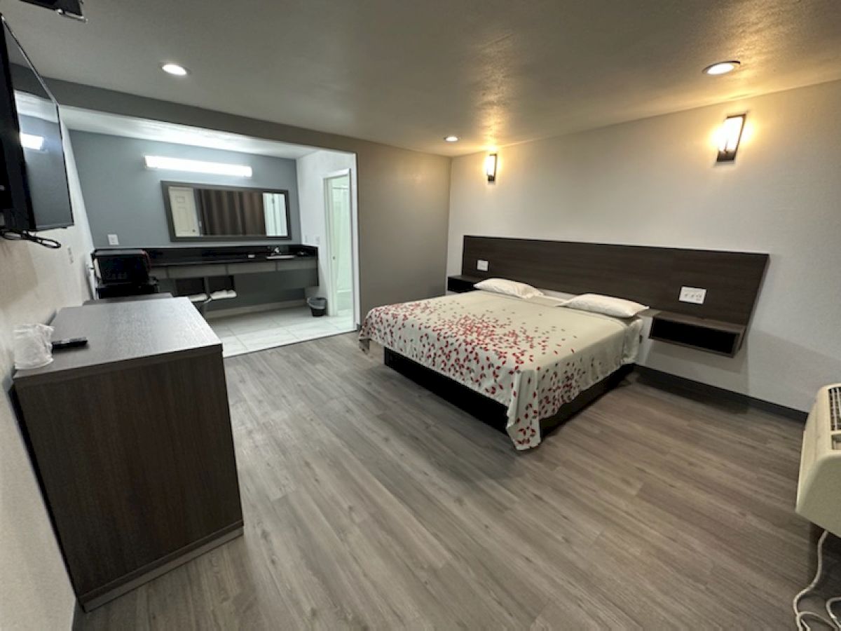 This image shows a modern hotel room featuring a bed, wall-mounted TV, wooden furniture, and a stylish bathroom with a vanity mirror.