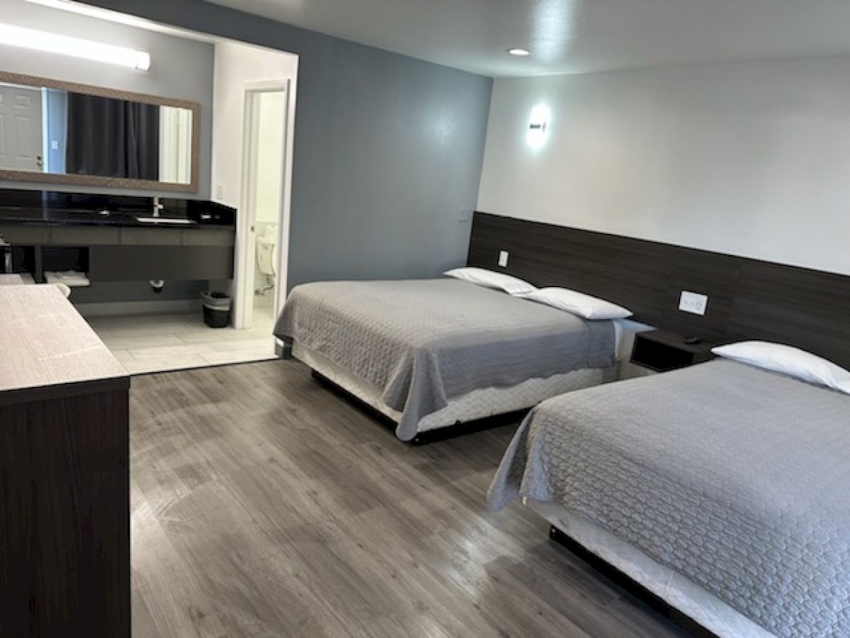 The image shows a modern hotel room with two beds, wood flooring, a wall-mounted light, a mirror, and a bathroom area at the back.