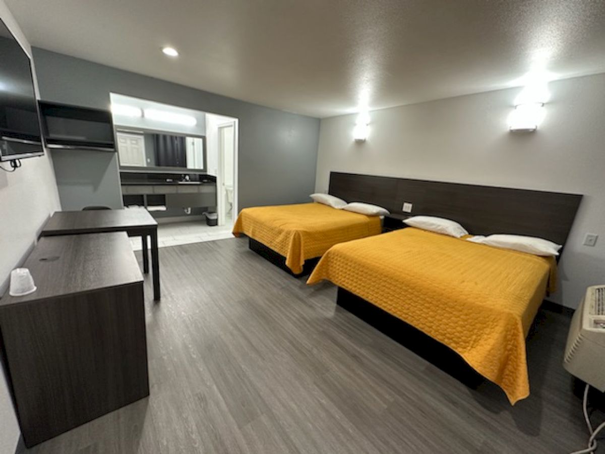 The image shows a hotel room with two double beds, yellow bedspreads, a desk, TV, and a view into a bathroom area.