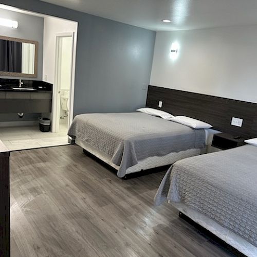 The image shows a clean, modern hotel room with two double beds, hardwood floors, and a bathroom in the background.