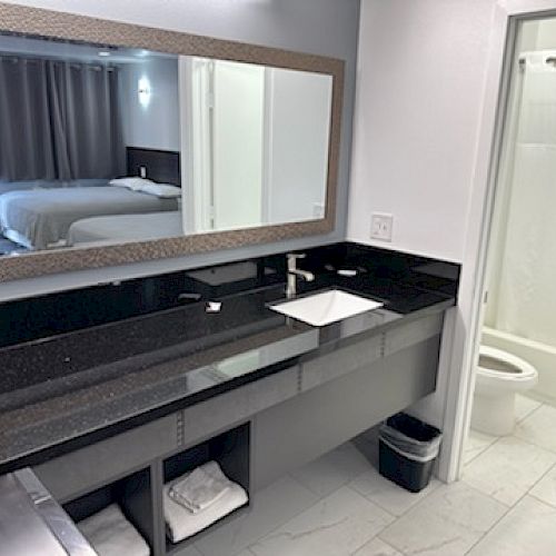 The image shows a hotel bathroom with a large mirror, black countertop, sink, and a view into a bedroom. A toilet and shower are visible.