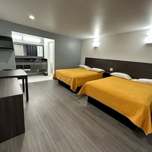 The image shows a hotel room with two beds with yellow covers, a TV, a desk, and a bathroom area in the background.